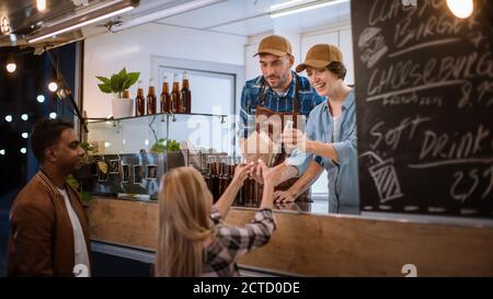 Mitarbeiter von Food Truck gibt Burger und kaltes Getränk an einen glücklichen jungen Hipster-Kunden weiter. Erfolgreiche Commercial Truck Verkauf Street Food in Modern Cool Stockfoto