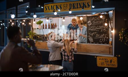Food Truck Mitarbeiter gibt frisch zubereitete Rinderburgers, Pommes und kalte Getränke an glückliche junge Hipster-Kunden. Commercial Truck Verkauf Street Food Stockfoto