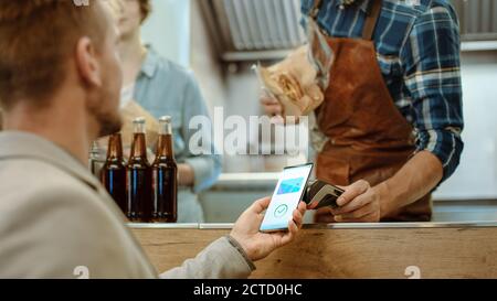 Food Truck Mitarbeiter gibt einen frisch gebackenen Burger an einen glücklichen jungen Mann. Mann in einem Casual Anzug verwendet sein Smartphone für NFC Mobile Payment Stockfoto