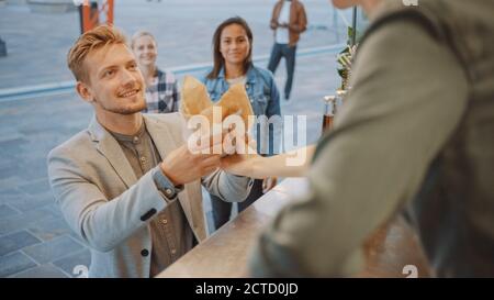 Mitarbeiter von Food Truck gibt einem glücklichen jungen Mann in einem Anzug frisch zubereitete Burger. Street Food Truck verkauft Burger in einem modernen Hip Nachbarschaft Stockfoto