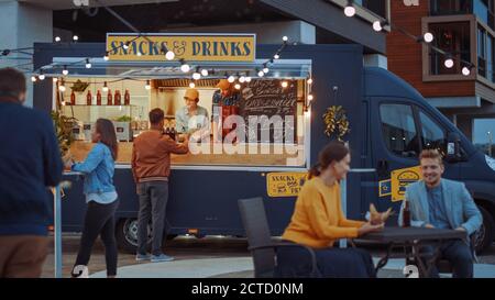 Mitarbeiter von Food Truck übergibt ein Kartenterminal für kontaktlose Zahlungen an den jungen Hipster-Kunden, der einen leckeren Burger kauft. Nutzfahrzeug Stockfoto