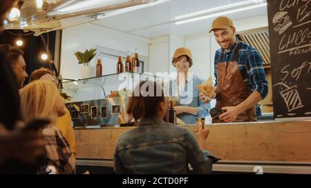 Food Truck Employee gibt einem glücklichen jungen Weibchen einen frisch gebackenen Burger. Junge Dame bezahlt für Essen mit kontaktloser Kreditkarte. Street Food Stockfoto