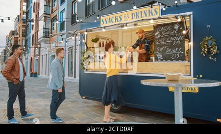 Food Truck Employee gibt einen frisch zubereiteten Gourmet Burger an glückliche junge Männer und Frauen in der Warteschlange. Street Food Truck Verkauf Burger im Freien in einem Stockfoto