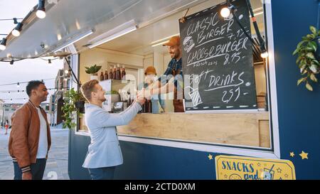 Food Truck Mitarbeiter gibt einem glücklichen jungen Mann in einem Anzug einen frisch zubereiteten Gourmet Burger. Street Food Truck Verkauf Burger in einem modernen Hip Stockfoto