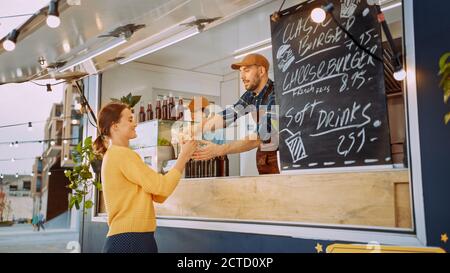 Food Truck Employee überreicht einen frisch zubereiteten Gourmet Burger an eine glückliche junge Frau. Der junge Mann in einem Anzug macht einen Auftrag. Street Food Truck Selling Stockfoto