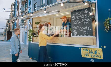 Food Truck Employee überreicht einen frisch zubereiteten Gourmet Burger an eine glückliche junge Frau. Der junge Mann in einem Anzug macht einen Auftrag. Street Food Truck Selling Stockfoto
