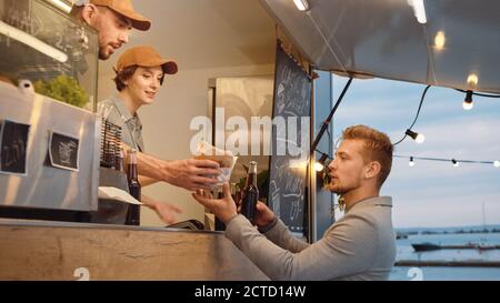 Food Truck Mitarbeiter gibt Rindfleisch Burger, Pommes und kalte Getränke an Happy Hipster Male. Die Leute essen draußen. Commercial Truck Selling Street Stockfoto