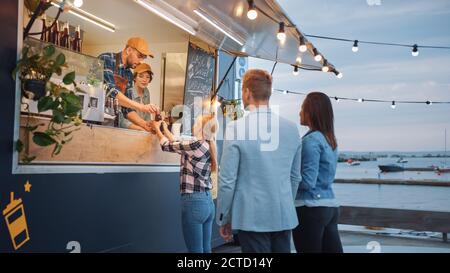 Food Truck Mitarbeiter gibt Rindfleisch Burger, Pommes und kalte Getränke an glückliche Hipster Kunden. Die Leute essen draußen. Verkauf Von Nutzfahrzeugen Stockfoto