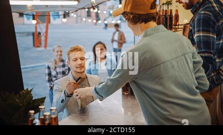 Mitarbeiter von Food Truck übergibt einen frisch zubereiteten Burger an Happy Young man in einem Anzug. Männlich bezahlt für Essen mit kontaktloser Kreditkarte. Street Food Stockfoto