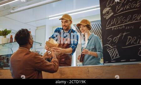 Mitarbeiter von Food Truck gibt Burger und kaltes Getränk an einen glücklichen jungen Hipster-Kunden weiter. Commercial Truck verkauft Street Food in moderner cooler Nachbarschaft Stockfoto