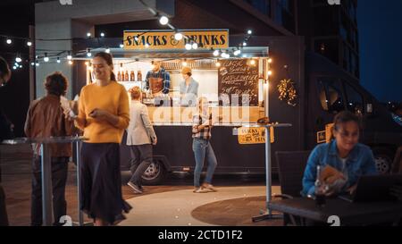 Food Truck Mitarbeiter gibt Rindfleisch Burger, Pommes und kalte Getränke an glückliche Hipster Kunden. Die Leute essen draußen an Tischen. Nutzfahrzeug Stockfoto