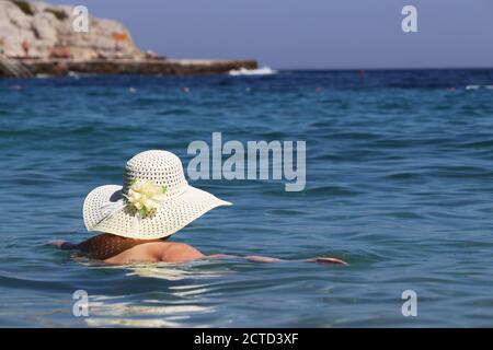 Frau in Sonnenhut schwimmen im blauen Meer auf Felsen Hintergrund. Entspannung auf dem Wasser, Strandurlaub