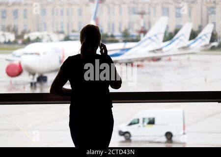 Silhouette einer Frau, die im Flughafenterminal auf dem Mobiltelefon spricht. Passagier wartet auf ihren Flug und schaut zu den Flugzeugen Stockfoto