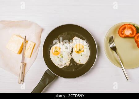 Draufsicht Bratpfanne mit zwei Spiegeleiern neben Butter, Messer und Teller mit Tomaten auf weißem Tisch. Stockfoto