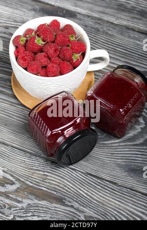 Himbeermarmelade in Gläsern. In der Nähe befinden sich Himbeeren in einem großen weißen Becher. Auf Holzbrettern mit einer schönen Textur. Stockfoto