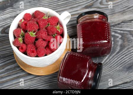 Himbeermarmelade in Gläsern. In der Nähe befinden sich Himbeeren in einem großen weißen Becher. Auf Holzbrettern mit einer schönen Textur. Stockfoto