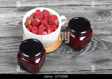 Himbeermarmelade in Gläsern. In der Nähe befinden sich Himbeeren in einem großen weißen Becher. Auf Holzbrettern mit einer schönen Textur. Stockfoto