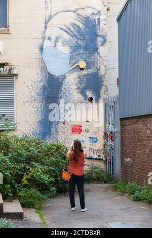 Weibliche Touristen fotografieren Banksy’s Straßenkunst „Girl with the Pierced Eardrum“. Während der Pandemie von Covid 19 hat sie eine Gesichtsmaske an. Bristol, Engl Stockfoto