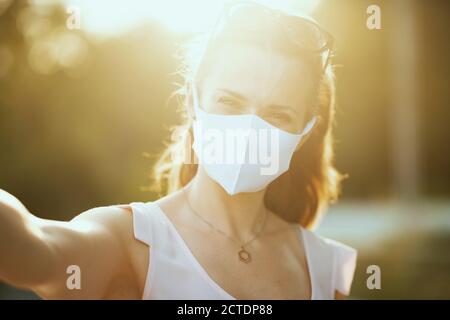 Porträt von glücklich elegante Frau in rosa Bluse mit weißer Maske und Sonnenbrille Selfie im Freien im Stadtpark. Stockfoto