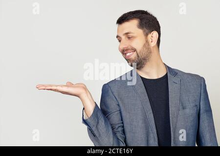 Junger Mann mit einem Bart in einem weißen T-Shirt lächelnd, fröhlich präsentiert und zeigt mit seiner Handfläche, Blick auf seine Hand. Steht auf isoliertem gelben Hintergrund. Stockfoto