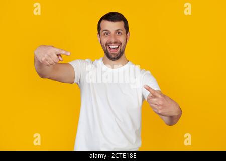 Junger Mann mit einem Bart in einem weißen T-Shirt, der mit den Fingern auf die Kamera zeigt, mit einem fröhlichen und lustigen Gesicht. Gute Energie und Vibes. Steht auf isoliertem gelben Hintergrund. Stockfoto