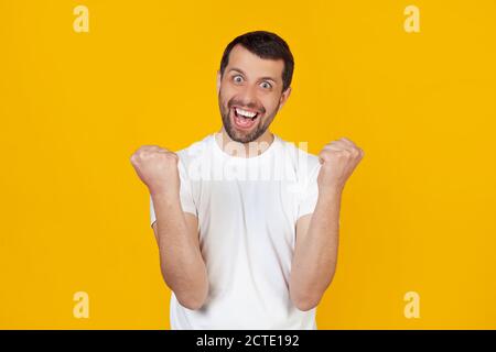 Ein junger Mann mit einem Bart in einem weißen T-Shirt ist sehr glücklich und aufgeregt, macht eine Gewinnergeste mit erhobenen Händen, lächelt und schreit Erfolg. Celebration Concept steht auf isoliertem gelben Hintergrund Stockfoto