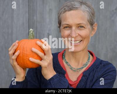 Die kaukasische kanadische Landsfrau mittleren Alters mit kurzen Haaren hält einen Kürbis in den Händen und lächelt an der Kamera. Stockfoto