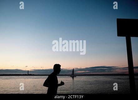 Ein Mann joggt an der Küste bei West Cowes entlang In der Dämmerung Abenddämmerung segelt eine Yacht vorbei auf der Solent am Strand raus zum Meer Stockfoto