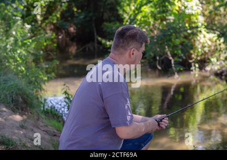 Ein erwachsener Mann fängt einen Raubfisch am Ufer eines Waldflusses. Ein kurzhaariger Fischer mit einer Rute in der Hand. Aktiver Lebensstil. Selektiver Fokus Stockfoto