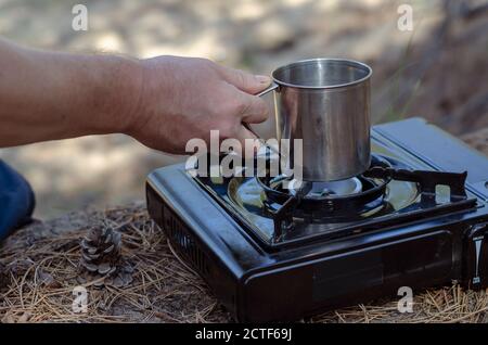 Kräutertee in einem Metallbecher in der Hand über einem tragbaren Gasherd. Eine Männerhand hält Kräutertee aus Hagebutten und verschiedenen Kräutern in einem Edelstahl Stockfoto