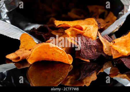 Beutel mit Gemüsechips auf schwarzem Hintergrund reflektiert. Geschmack von Süßkartoffeln, Rote Beete und Pastinak. Veganes Lebensmittelkonzept Stockfoto