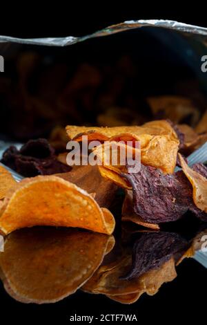 Beutel mit Gemüsechips auf schwarzem Hintergrund reflektiert. Geschmack von Süßkartoffeln, Rote Beete und Pastinak. Veganes Lebensmittelkonzept Stockfoto