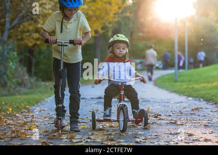 Schöne blonde zwei Jahre alten Kleinkind Junge und sein älterer Bruder, Reiten roten Dreirad und Roller im Park bei Sonnenuntergang, schönen Herbsttag Stockfoto