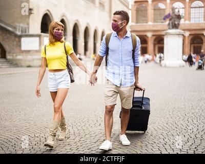 Ein glückliches Paar von Touristen, die eine Maske tragen, um sich vor Covid-19 zu schützen, laufen in der Stadt und halten Hände und Koffer. Neues normales Reisekonzept. Stockfoto