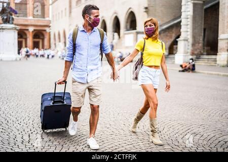 Ein glückliches Paar von Touristen, die eine Maske tragen, um sich vor Covid-19 zu schützen, laufen in der Stadt und halten Hände und Koffer. Neues normales Reisekonzept. Stockfoto