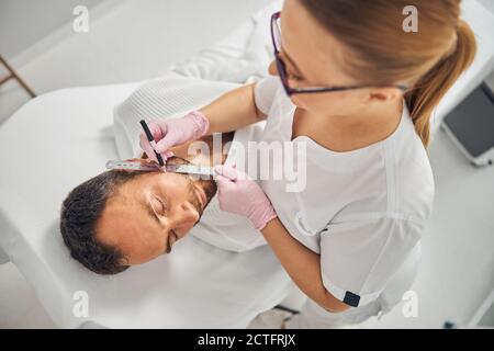 Kosmetikerin in sterilen Handschuhen platzieren Lineal auf jungen Mann Wange Und gerade Linie mit weißem Bleistift zeichnen Stockfoto