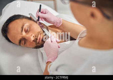 Close up of beautician in sterile gloves placing ruler on male cheek and drawing straight line with white pencil Stockfoto