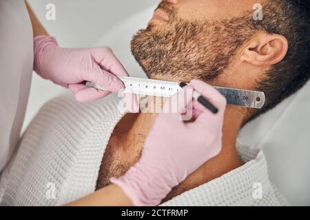 Nahaufnahme der Frau Hände in sterilen Handschuhen Platzierung Lineal Am männlichen Hals und Zeichnung gerade Linie mit weißem Bleistift Stockfoto