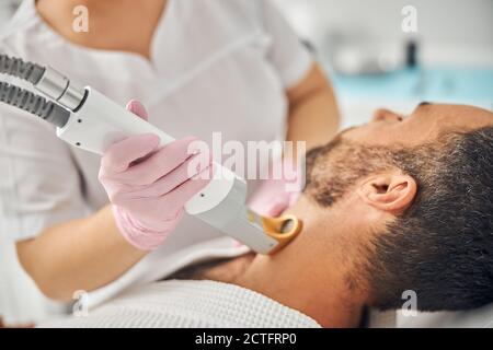 Weibliche Ästhetikerin Hände in sterilen Handschuhen entfernen unerwünschte Haare aus Männlicher Hals mit speziellem Lasergerät Stockfoto