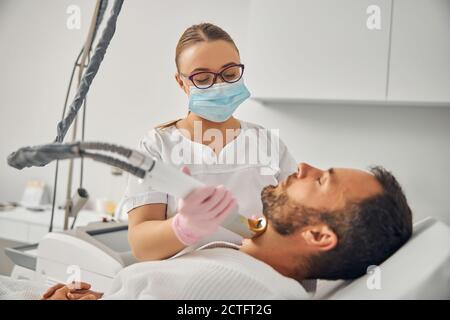 Charmante weibliche Kosmetikerin in medizinische Maske entfernen unerwünschte Haare aus Männlicher Hals mit Lasergerät Stockfoto