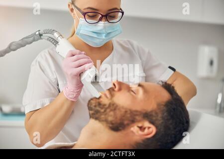 Bärtiger junger Mann, der auf dem Tagesbett liegt, während eine weibliche Kosmetologin entfernt Unerwünschte Haare aus der Wange mit Lasergerät Stockfoto