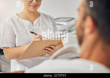 Lächelnder Arzt Kosmetologe schreiben auf Zwischenablage, während neben männlich sitzen Patient im Kosmetikschrank Stockfoto