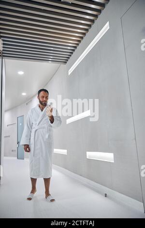 Bärtiger Herr im Bademantel hält Handy und lächelt im Stehen Im Korridor des modernen Wellness-Centers Stockfoto