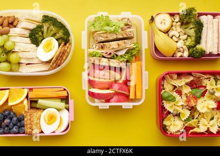 Draufsicht auf Schullunchboxen mit verschiedenen gesunden, nahrhaften Mahlzeiten auf gelbem Hintergrund. Stockfoto