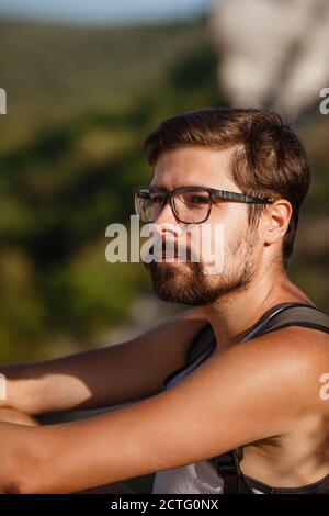 Junger männlicher Wanderer mit Rucksack, der sich während des ruhigen Sonnenuntergangs auf einem Berg entspannt - Landschaft aus dem Urlaub. Müder Reisender Mann, der auf einem Hügel ruht. Stockfoto