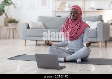 Yoga online. Schwarze muslimische Frau meditiert vor dem Laptop zu Hause Stockfoto