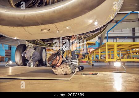 Rückansicht Porträt des Flugingenieurs auf dem Rücken liegend Unter dem offenen Motor während der Befestigung und Überprüfung des Systems von Passagierflugzeug im Hangar Stockfoto