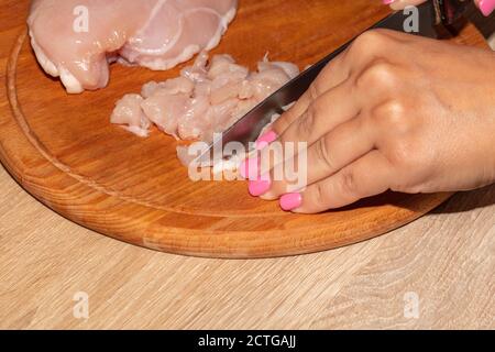 Eine Nahaufnahme von weiblichen Händen, die Hühnchen auf einem Holzbrett in Stücke schneiden. Pinke Fingernägel. Stockfoto