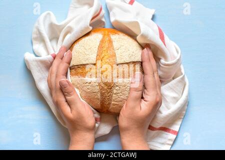 Weibliche Hände halten frisch gebackenes Sauerteig Brot in Serviette aus Ofen auf blauem Holztisch Draufsicht Flat Lay hausgemacht Gebäck Stockfoto