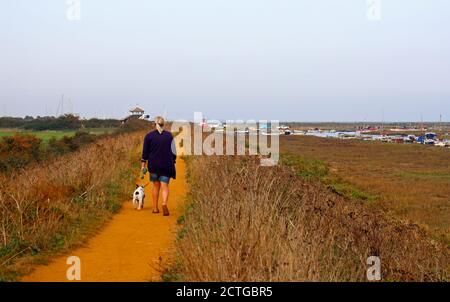 Eine Dame, die einen Hund auf dem Norfolk Coast Path von Morston Creek und den Salzwiesen in North Norfolk in Morston, Norfolk, England, Vereinigtes Königreich, läuft. Stockfoto